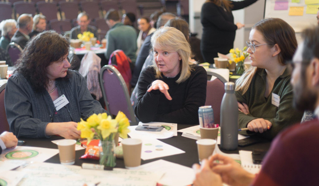 This picture shows people talking around a table at an event. It was taken by Alice Louisa